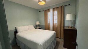 Une chambre avec un lit simple, une table de chevet avec une lampe, une commode en bois et une fenêtre avec des rideaux.