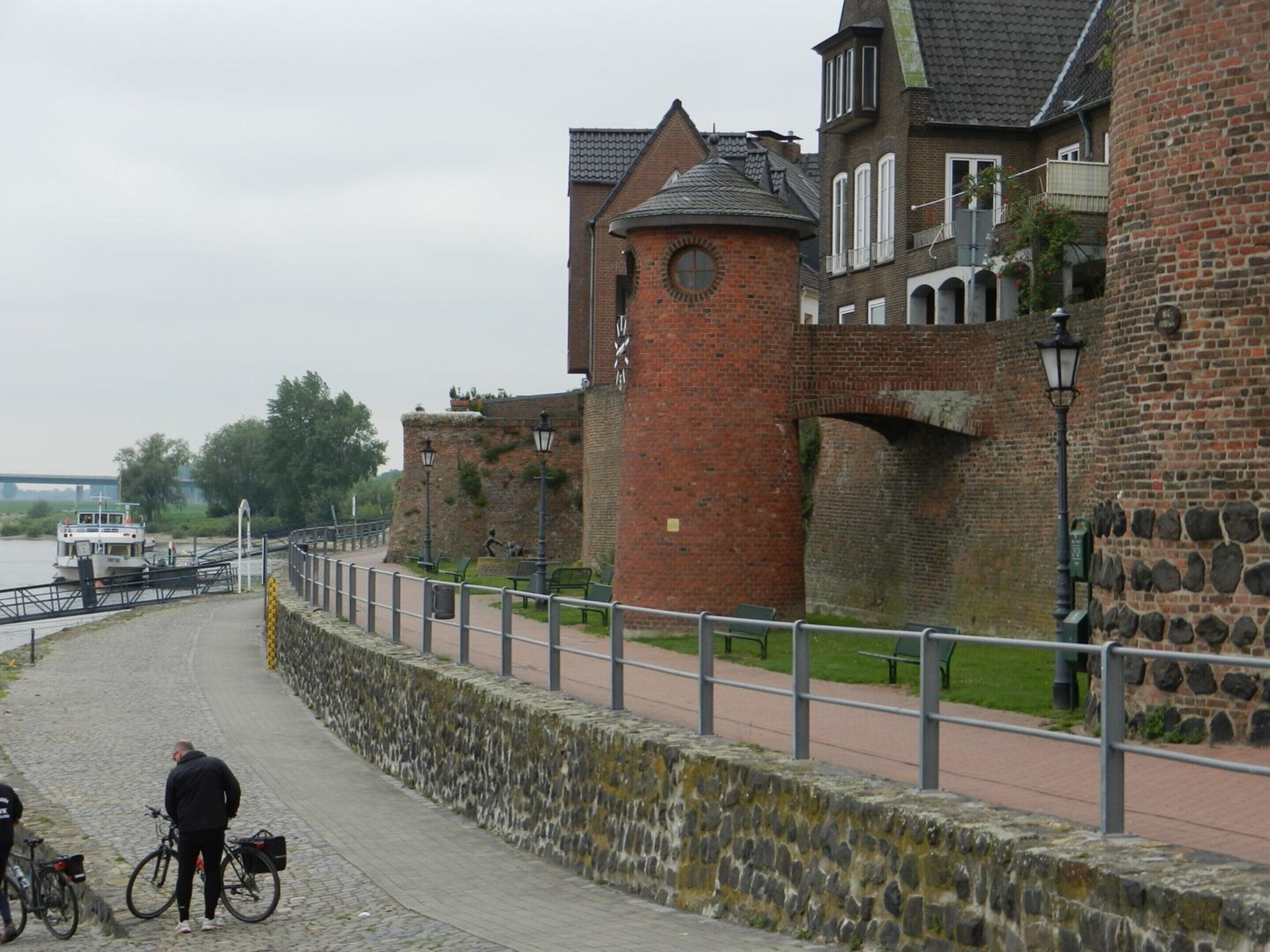 A historic brick building with a tower, a stone wall, and a paved walkway.
