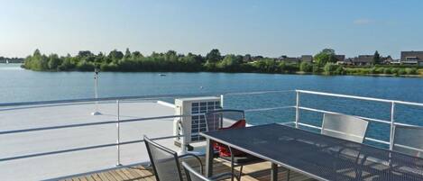 A deck with a table and chairs overlooking a calm body of water, with a view of a shoreline and buildings in the distance.