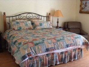 Un lit avec une couvre-lit à motifs et des oreillers, une tête de lit en bois, une lampe de chevet et une chaise, dans une pièce dont le sol est recouvert de parquet.