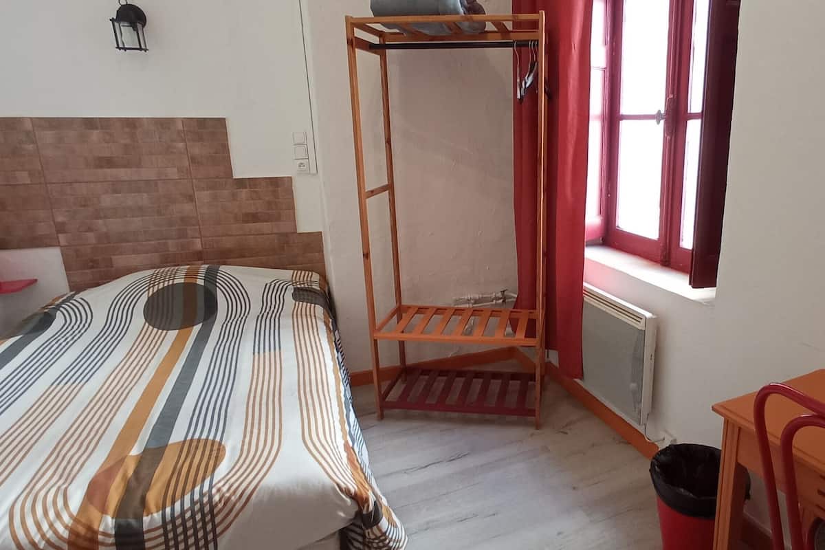 Une petite pièce bien éclairée, avec un lit, une étagère en bois, un radiateur et une fenêtre avec des rideaux.