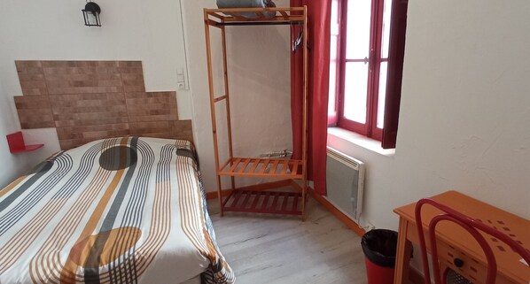 A small, well-lit room with a bed, a wooden shelf, a radiator, and a window with curtains.