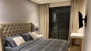 Een slaapkamer met een gestoffeerd hoofdeinde, een bed met een patroon, een nachtkastje met een lamp, een groot raam met jaloezieën, en een klein nachtkastje met een spiegel.