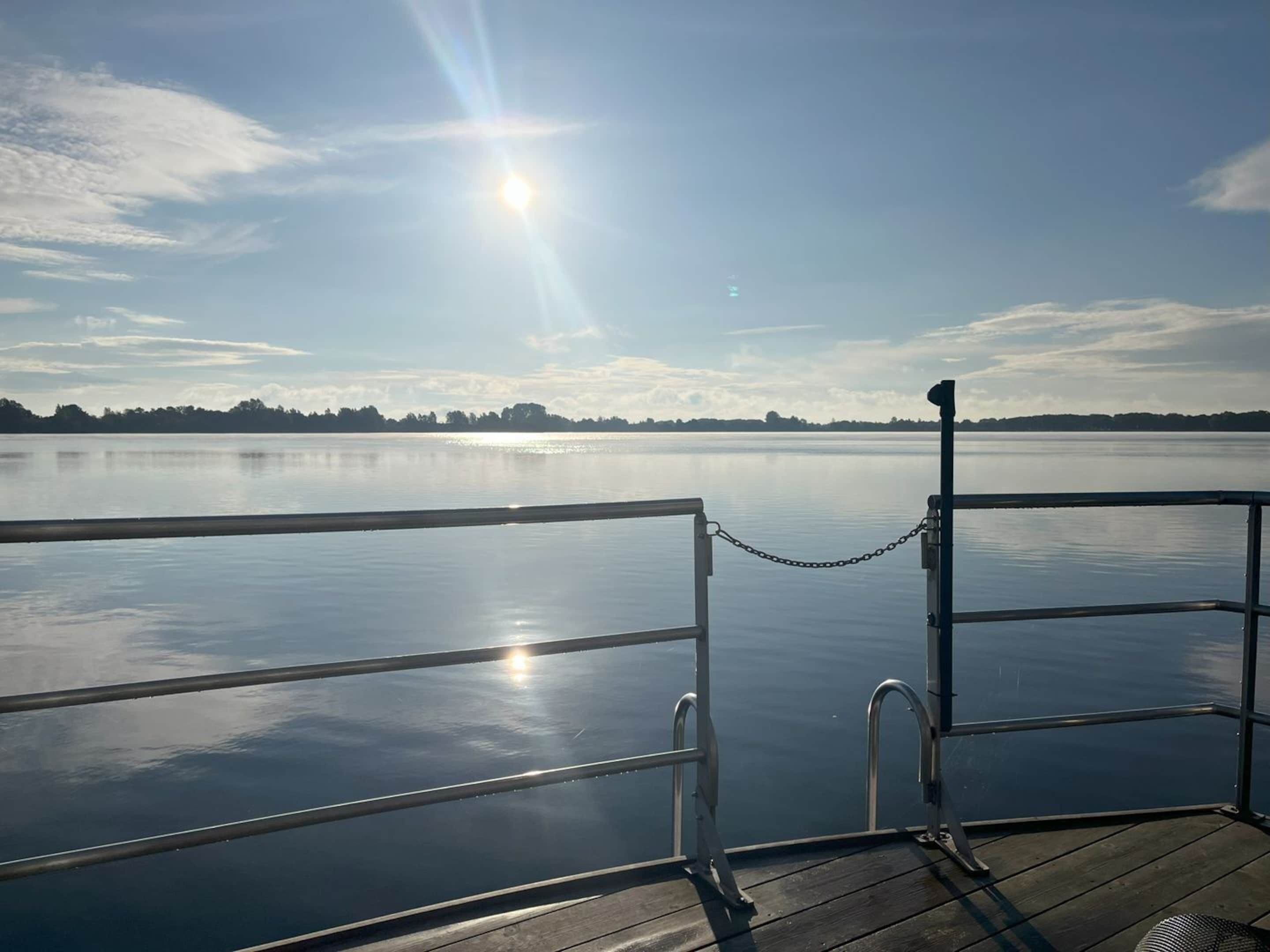 A wooden dock with metal railings and a chain barrier, overlooking a calm lake with a clear sky and distant trees.