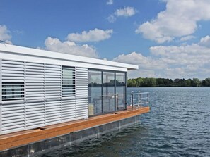 Een modern drijvend huis met een houten terras, grote ramen en uitzicht op een meer en bomen.