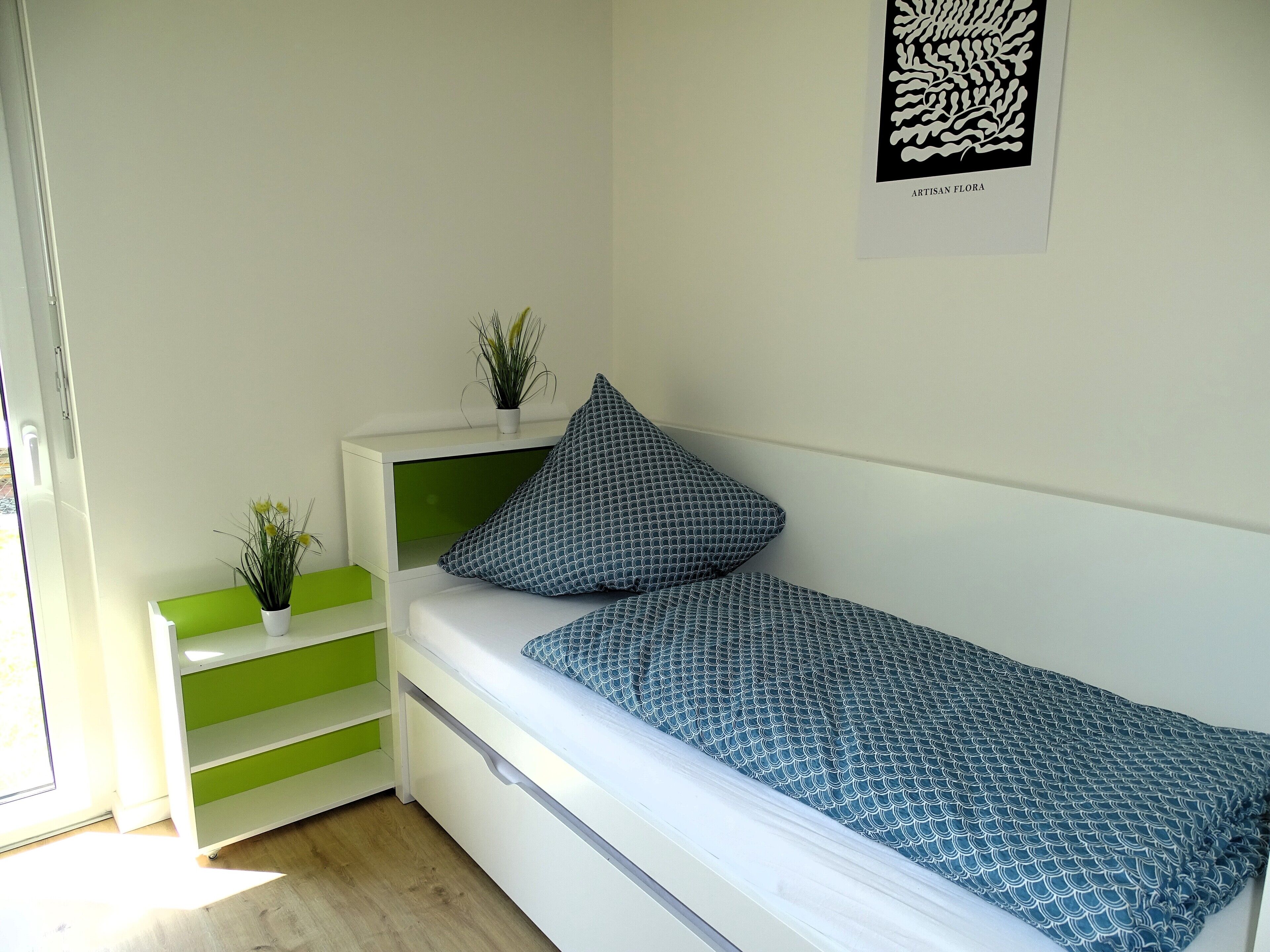 A neatly arranged bedroom with a bed, a bedside table with a plant, and a green storage unit.