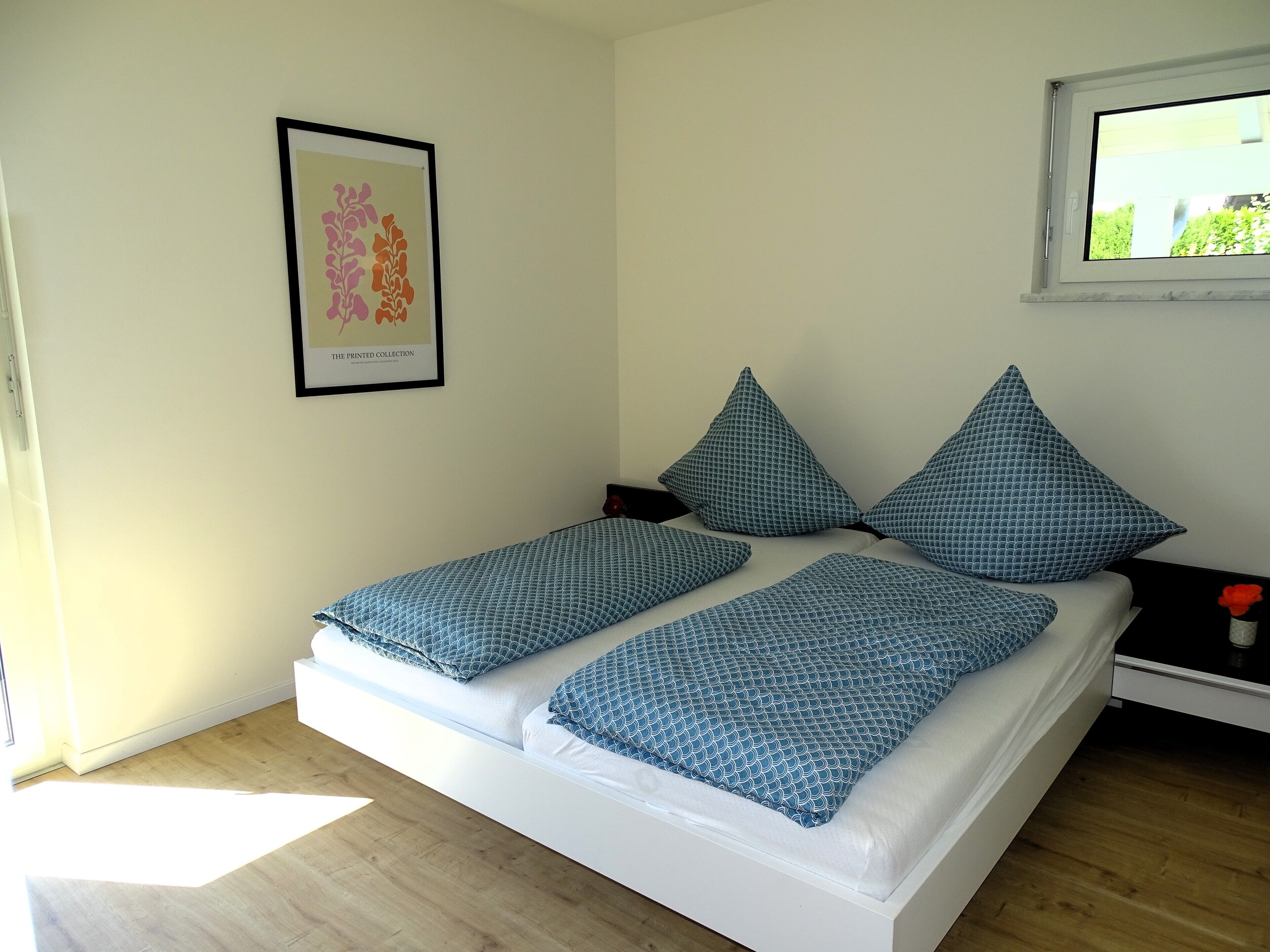 Une chambre à coucher avec un lit, deux oreillers, une table de chevet, un tableau encadré au mur et une fenêtre.