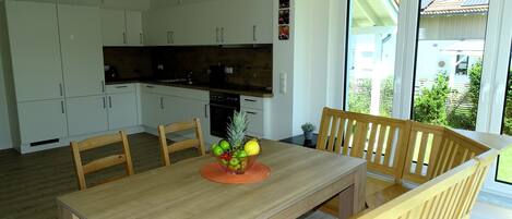 A modern kitchen with white cabinets, a wooden dining table with chairs, and a view of the outdoors through large windows.