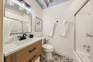 Features a sleek vanity, large mirror, and framed artwork, alongside a toilet and glass-enclosed shower.