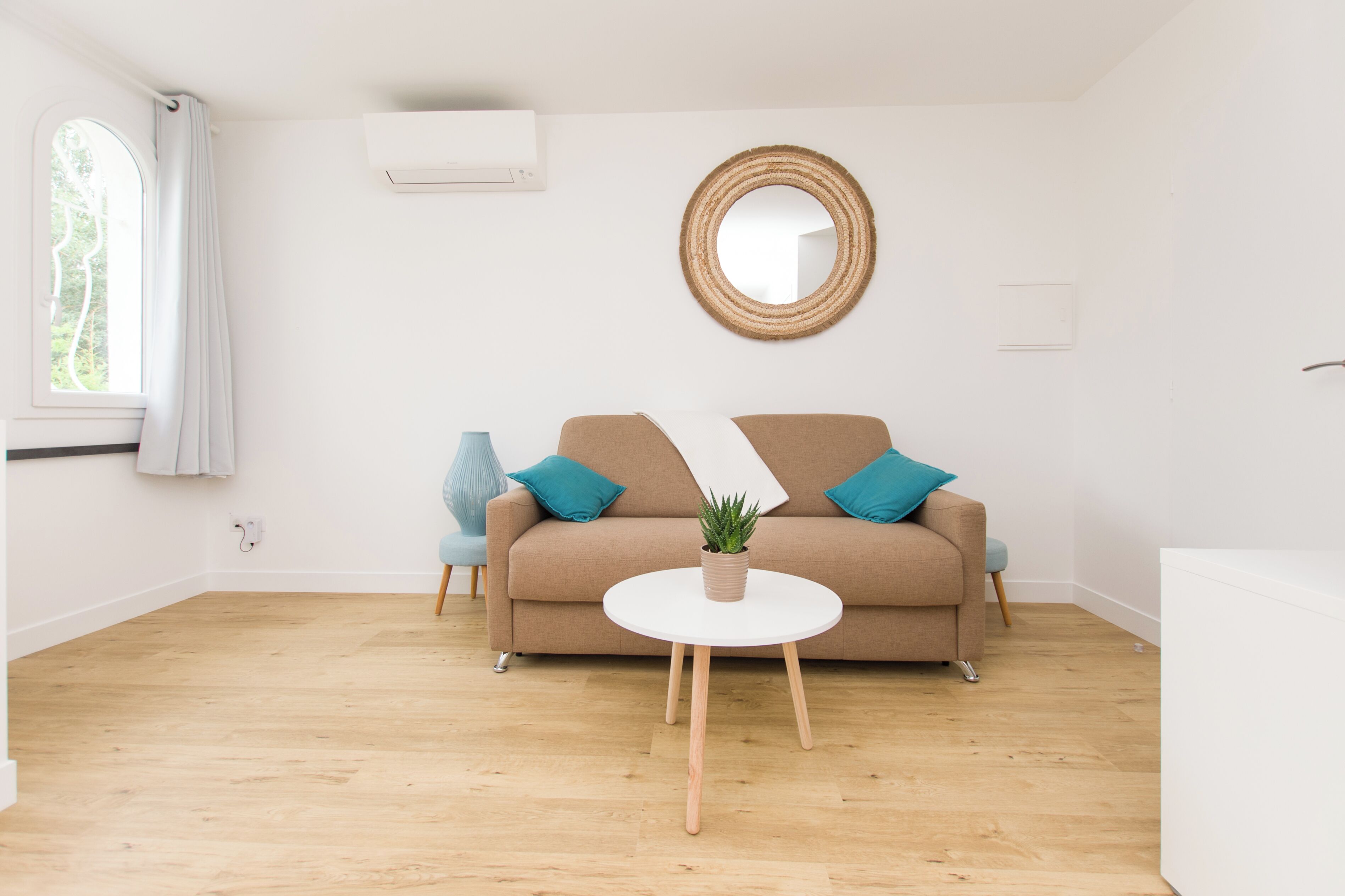 Una sala de estar moderna con un sofá, un espejo redondo, una mesita con una planta y una ventana con cortinas.