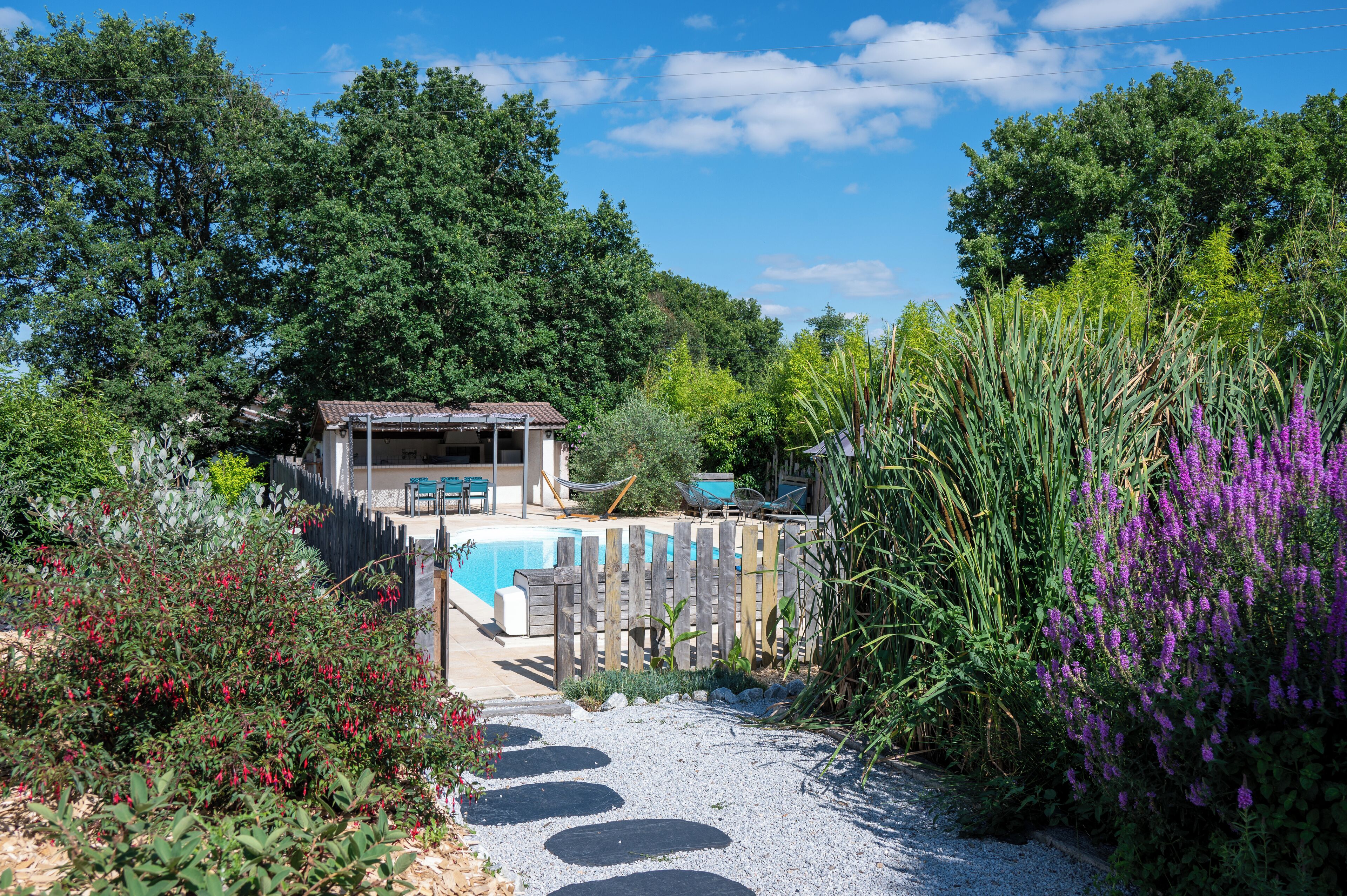 Un espace piscine doté d’une terrasse en bois, entouré d’une végétation luxuriante et de fleurs violettes.