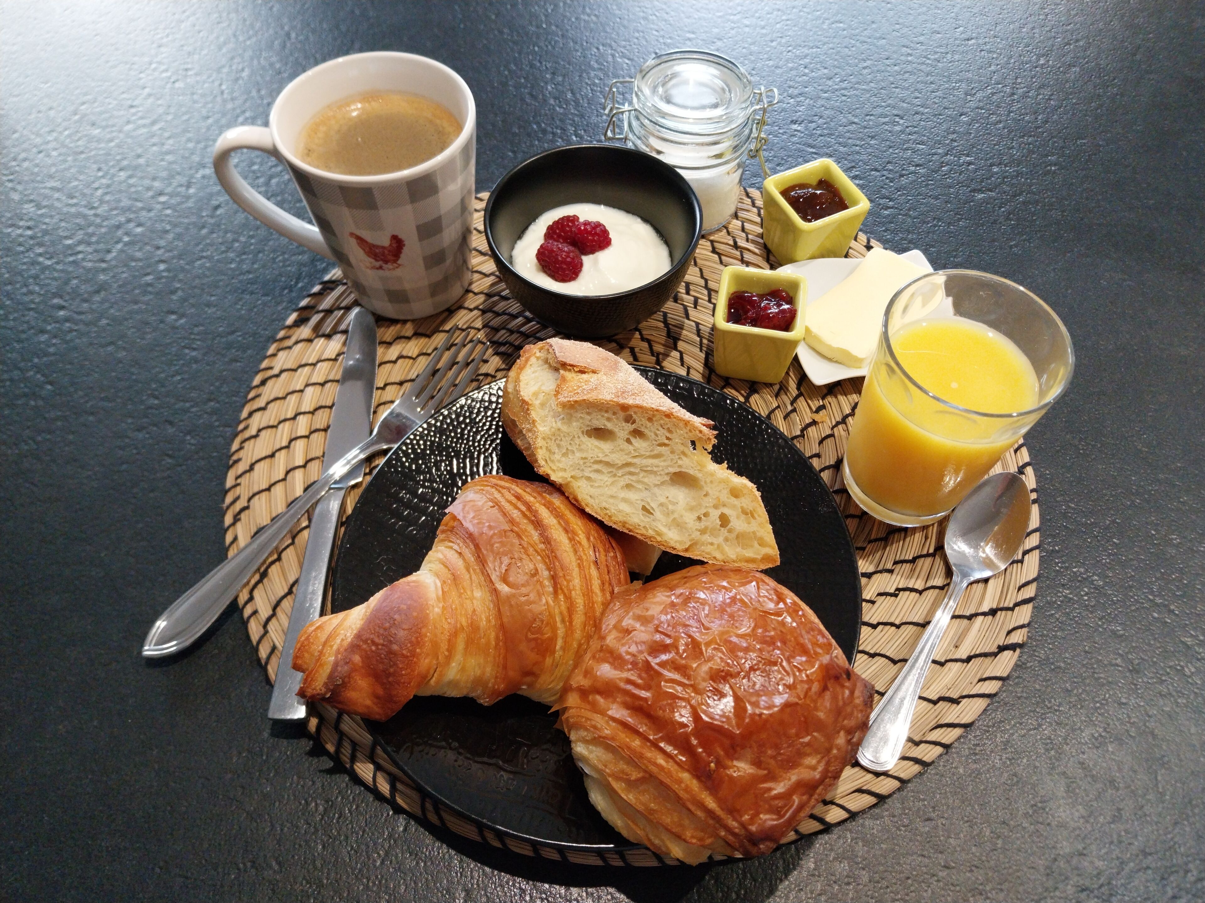 Une table dressée pour le petit-déjeuner avec des croissants, une tasse de café, du yaourt aux framboises, de la confiture, du fromage et du jus d’orange.
