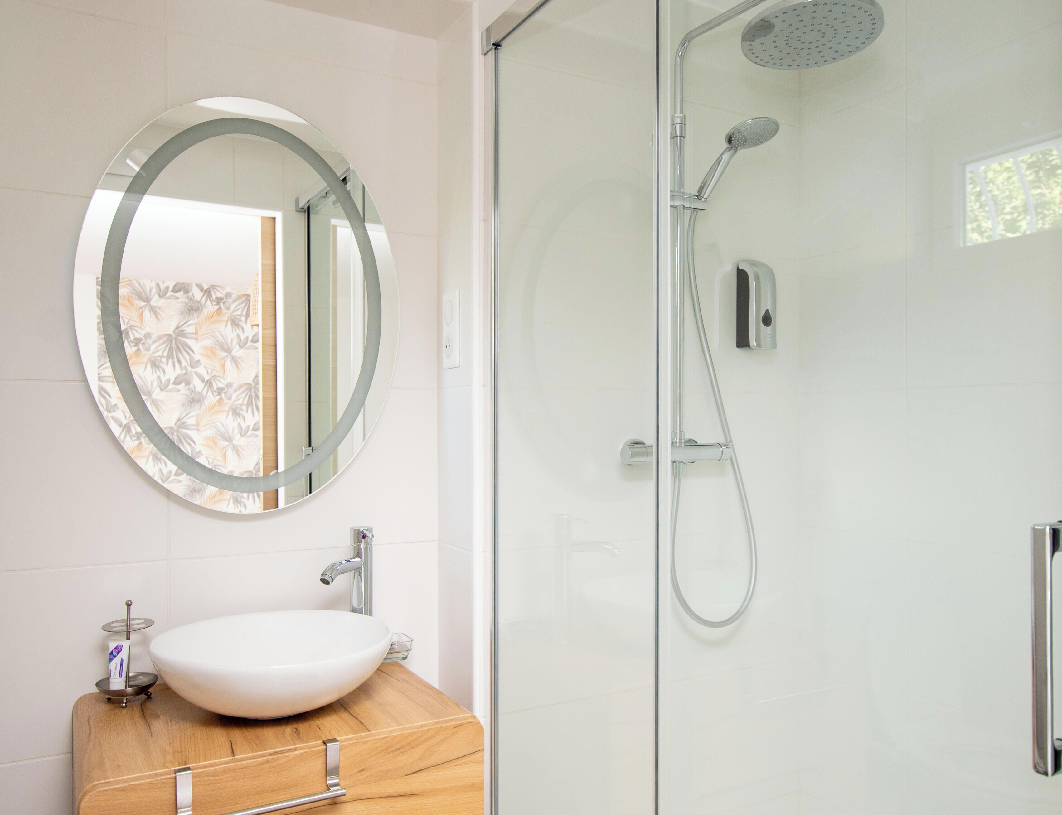 Un baño con un espejo redondo, un tocador de madera, un lavabo blanco y una zona de ducha con mampara de vidrio.