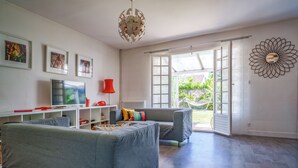 A modern living room with a grey sofa, a television, and a red lamp. There are framed pictures on the wall and a large window with a view of greenery outside. - Maison deco années 70 / 5 p. garden south Tours (Joué-lès-Tours)