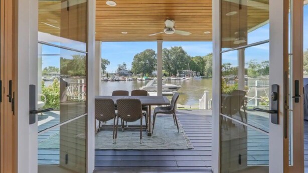 Un área de comedor exterior moderna con una mesa, sillas, ventilador de techo y vistas a un lago con barcos.
