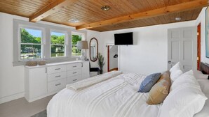 Un dormitorio con techo de madera, paredes blancas, una cama grande con ropa de cama blanca, un aparador, un espejo y un televisor montado en la pared.