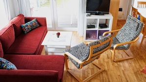 A living room with a red sofa, a patterned armchair, a small white table with a plant, and a TV on a white stand. - Hausboot in Barth am Barther Bodden (Barth)