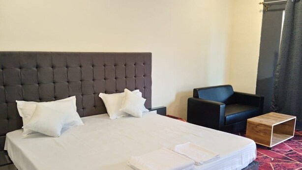 A neatly made bed with white linens and pillows, a dark upholstered headboard, a black leather chair, and a small wooden side table. - RASS Resort (Karjat)