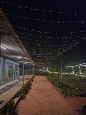 A pathway under a string-lit canopy, leading to a building with a blue exterior and a person walking towards the entrance. - RASS Resort (Karjat)