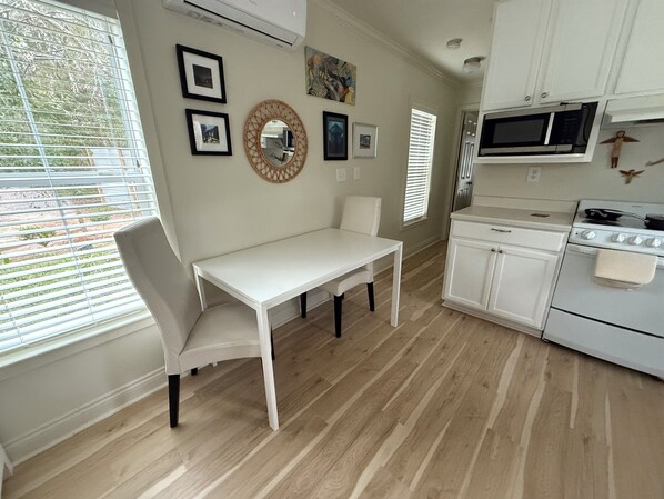 Una cocina con armarios blancos, una mesa de comedor blanca y una silla blanca. Hay cuadros en la pared, un microondas y una estufa.