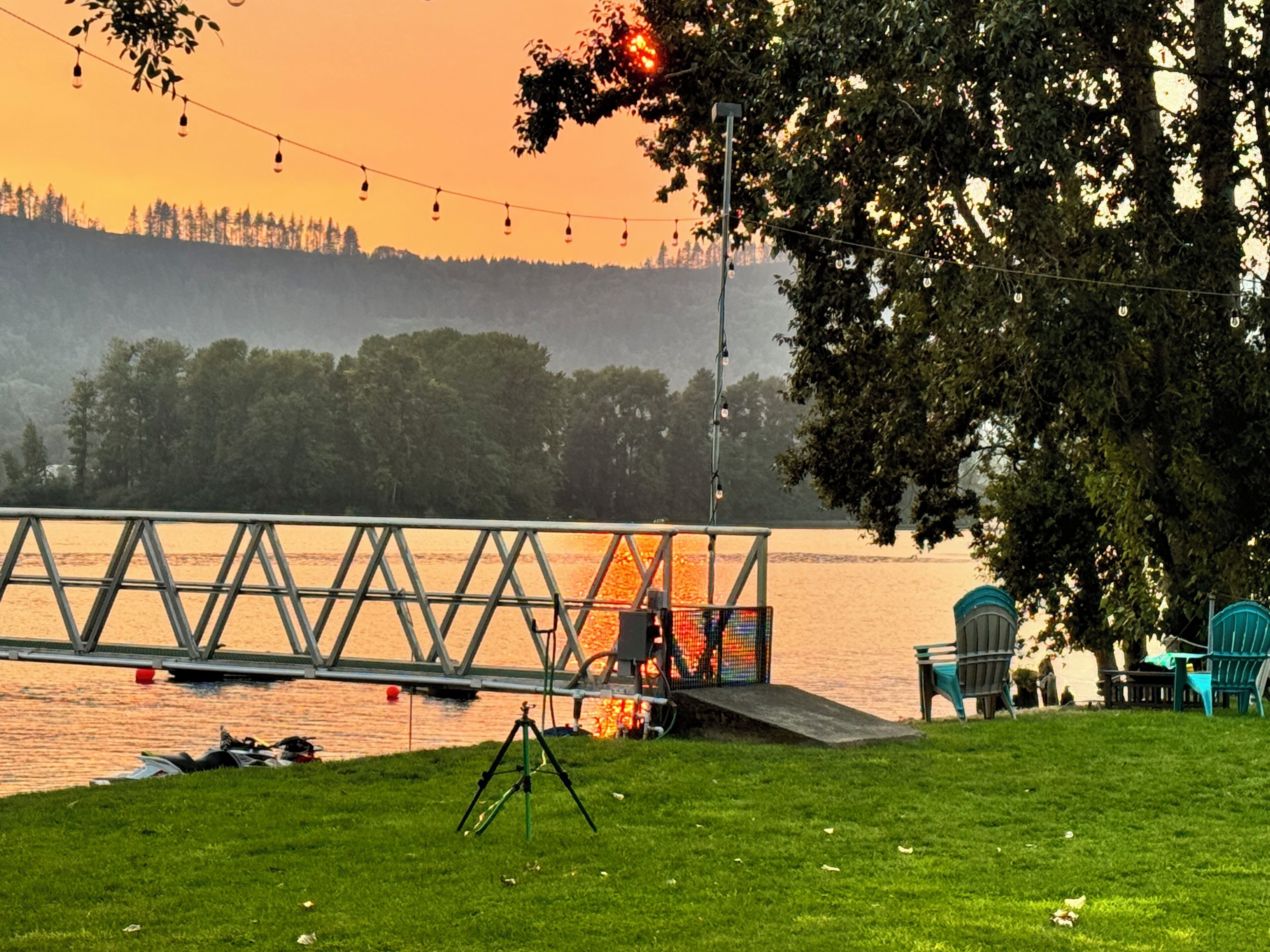 A lakeside view with a dock, chairs, and a tripod.