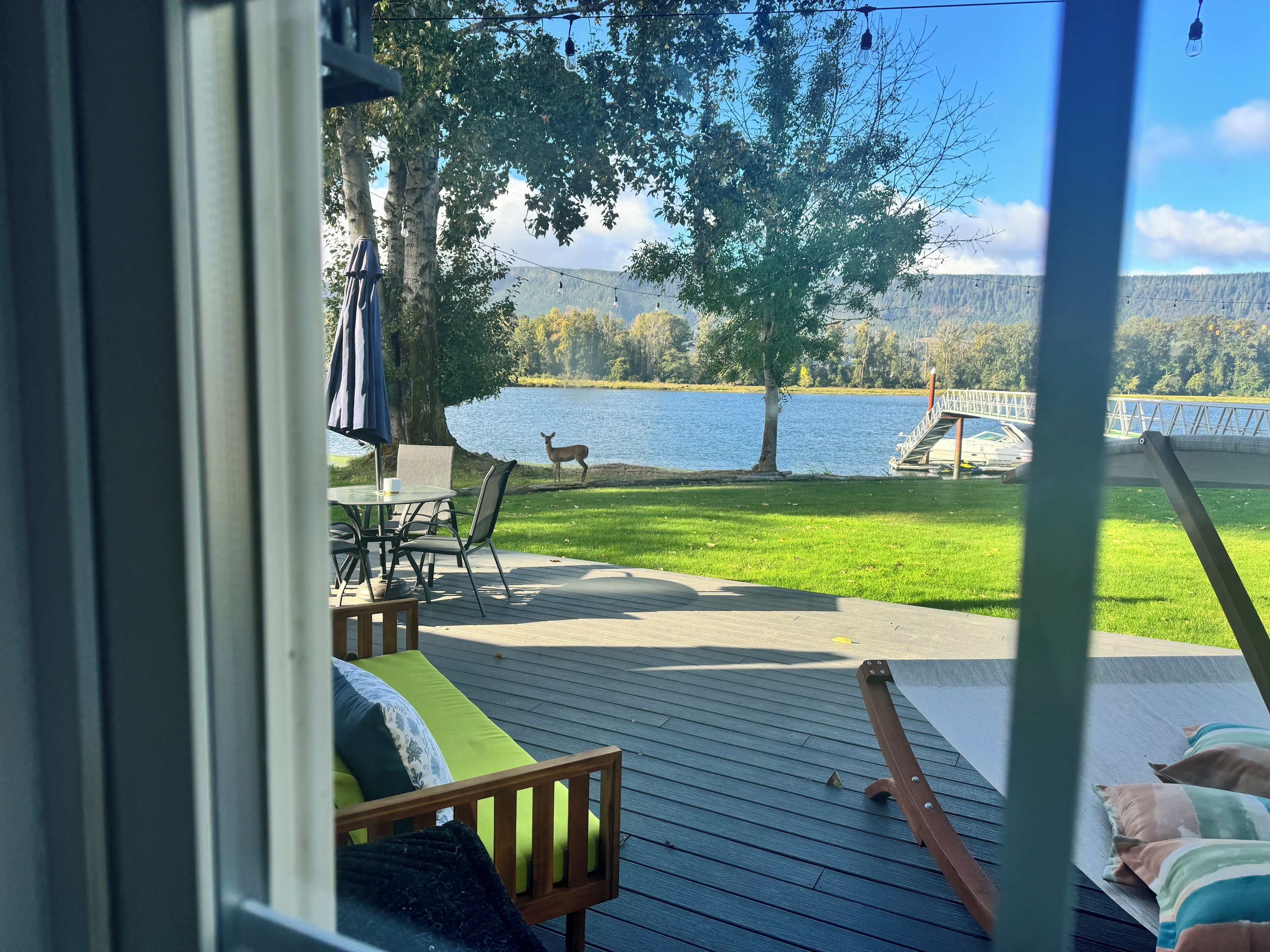 A deck with outdoor seating, a view of a lake, a deer, and a bridge.