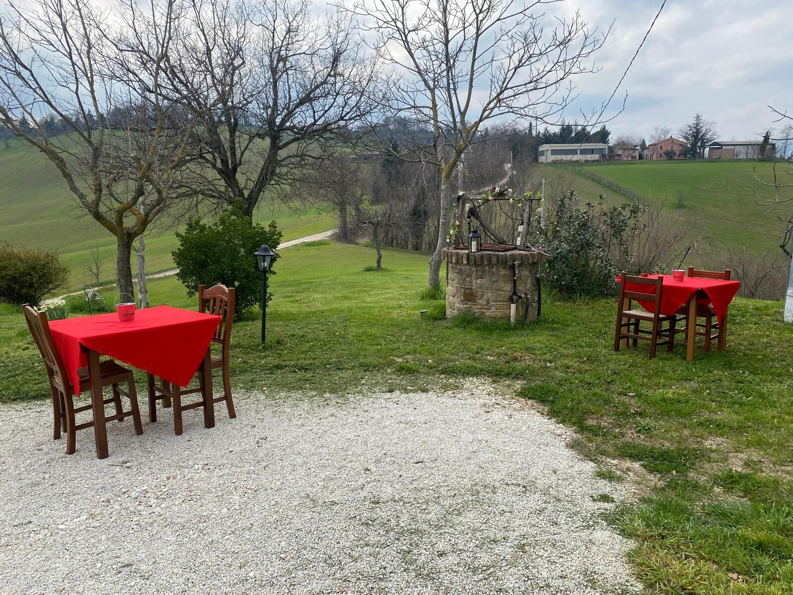 Un patio de grava con dos mesas y sillas de tejas rojas, rodeado de árboles y un paisaje cubierto de césped.