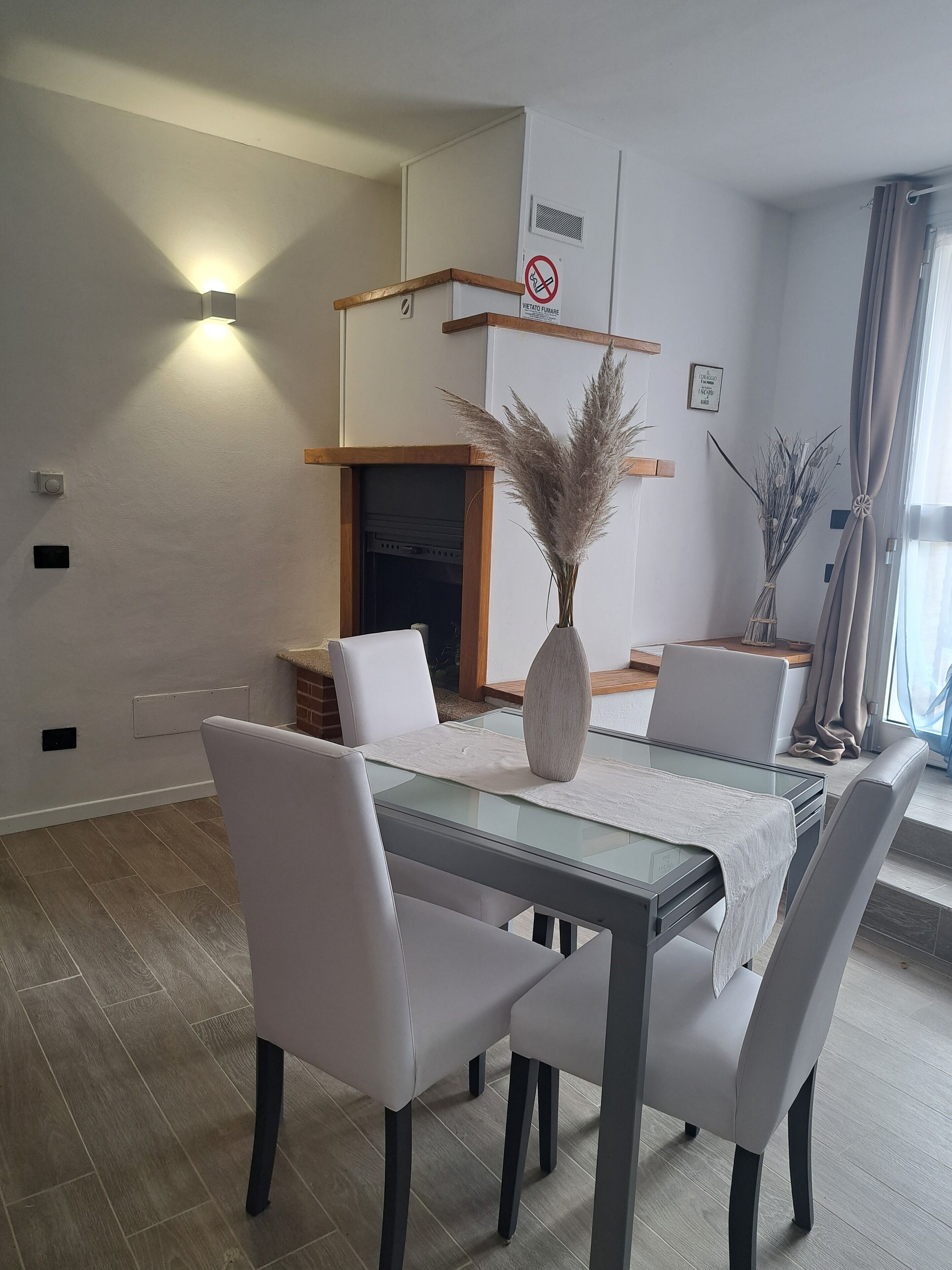 A modern dining area with a glass table, white chairs, a vase with pampas grass, and a fireplace.