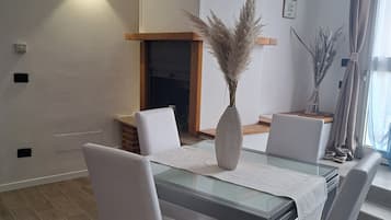 A modern dining area with a glass table, white chairs, a vase with pampas grass, and a fireplace.