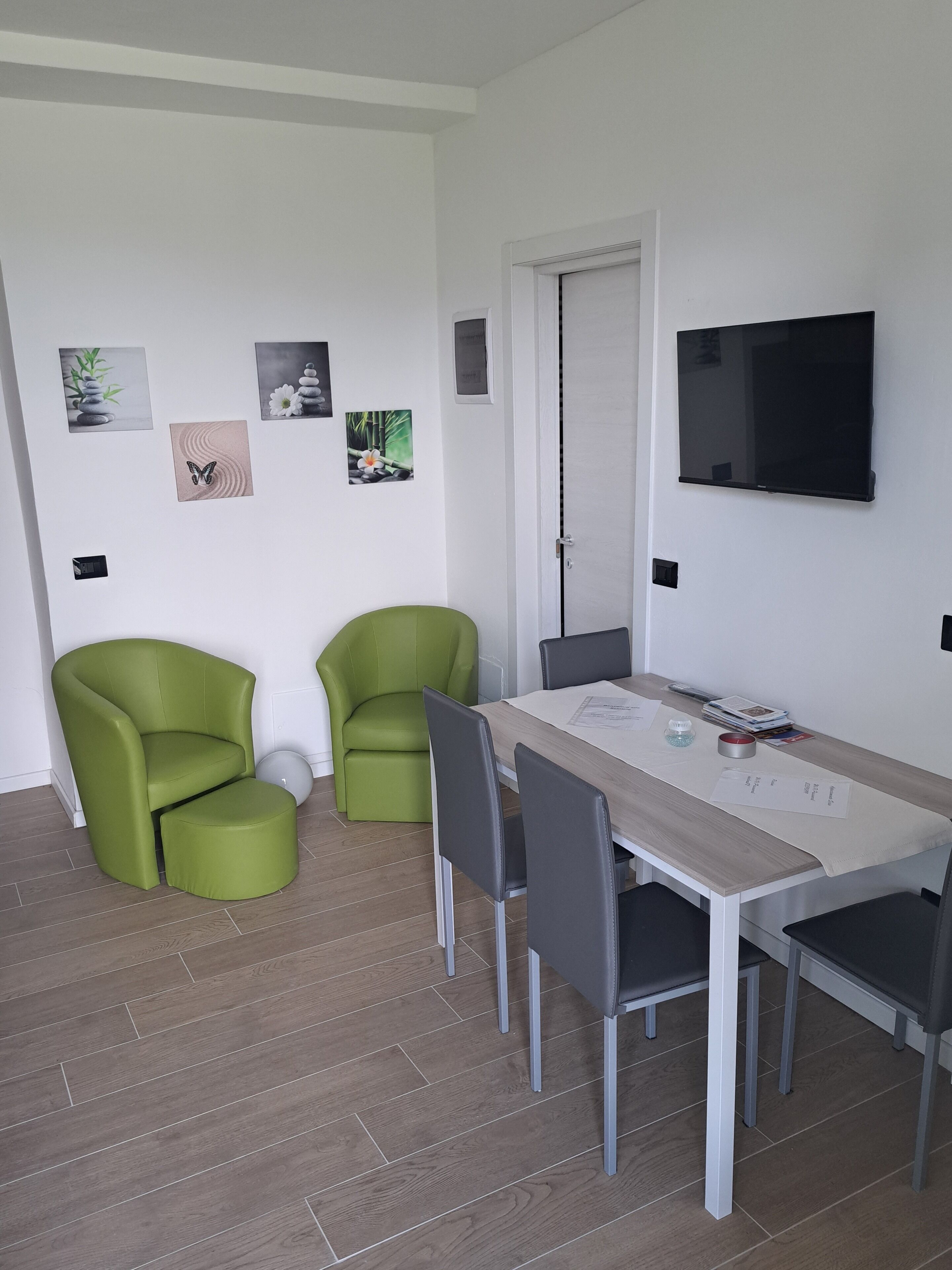 Una habitación moderna y luminosa con una mesa de comedor, dos sillas verdes, un televisor montado en la pared y una puerta.