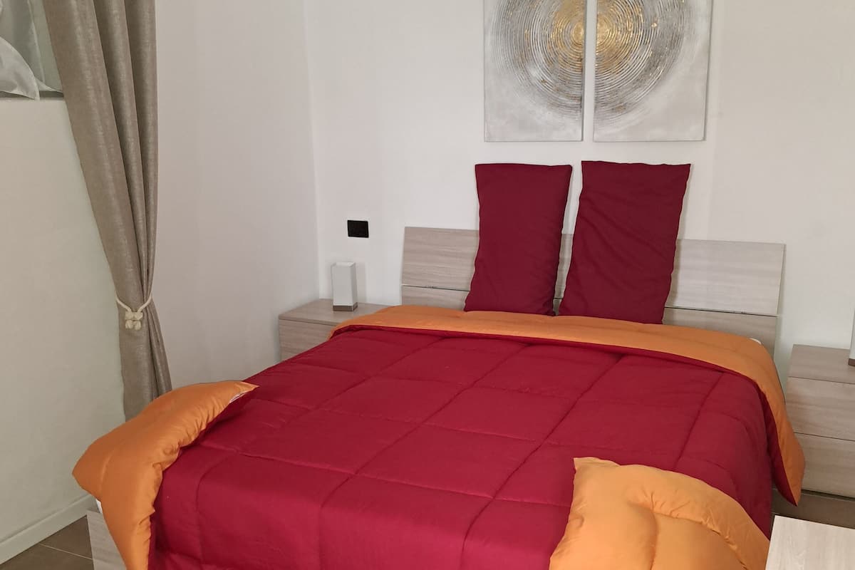 A bed with a red and orange bedspread, two red pillows, and a white pillow on a wooden frame. There is a white wall with a circular abstract painting above the bed.