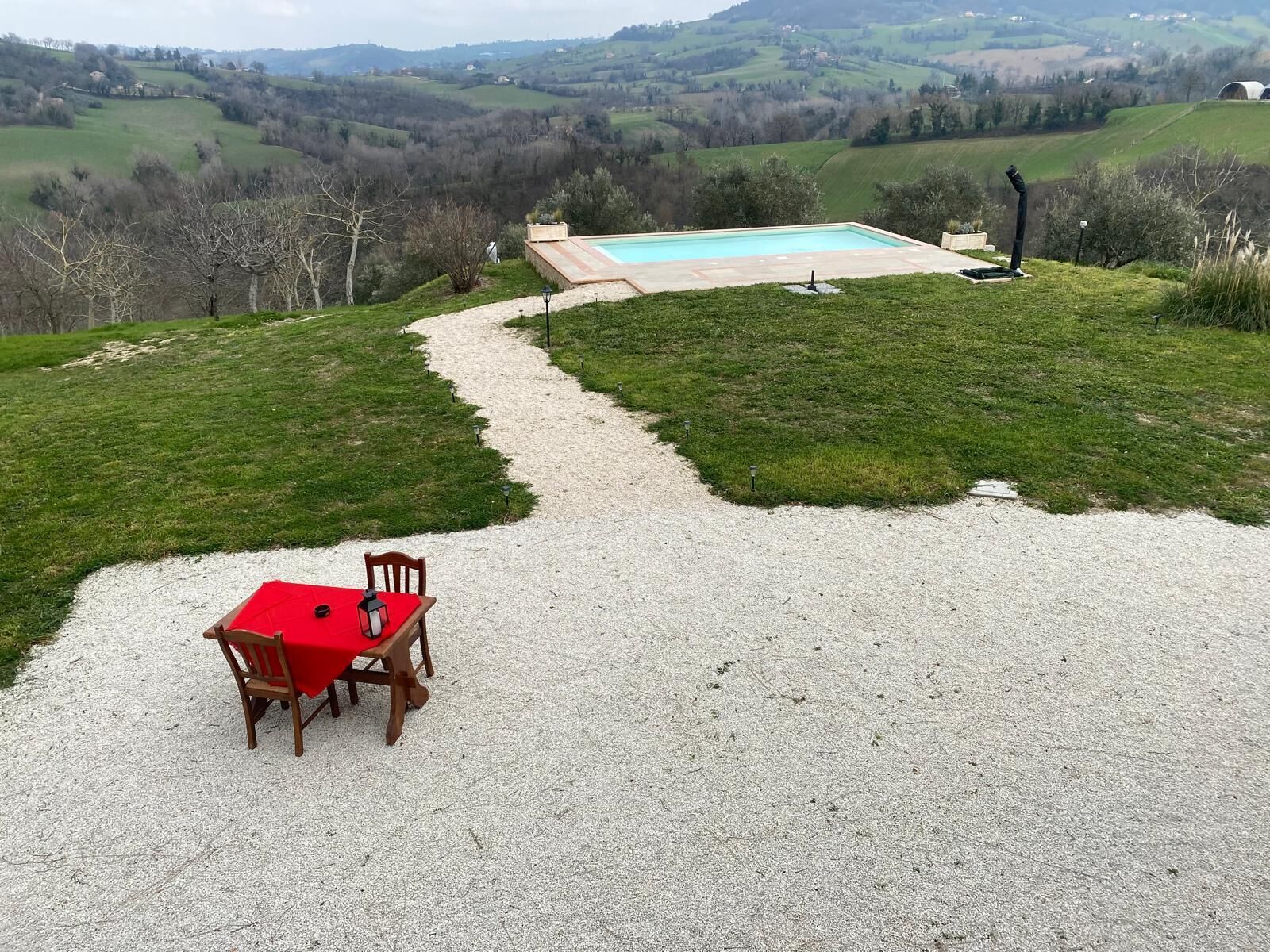 Une terrasse en gravier avec une table et des chaises rouges, qui mène à une piscine entourée d’une pelouse et offrant une vue sur des collines ondoyantes.