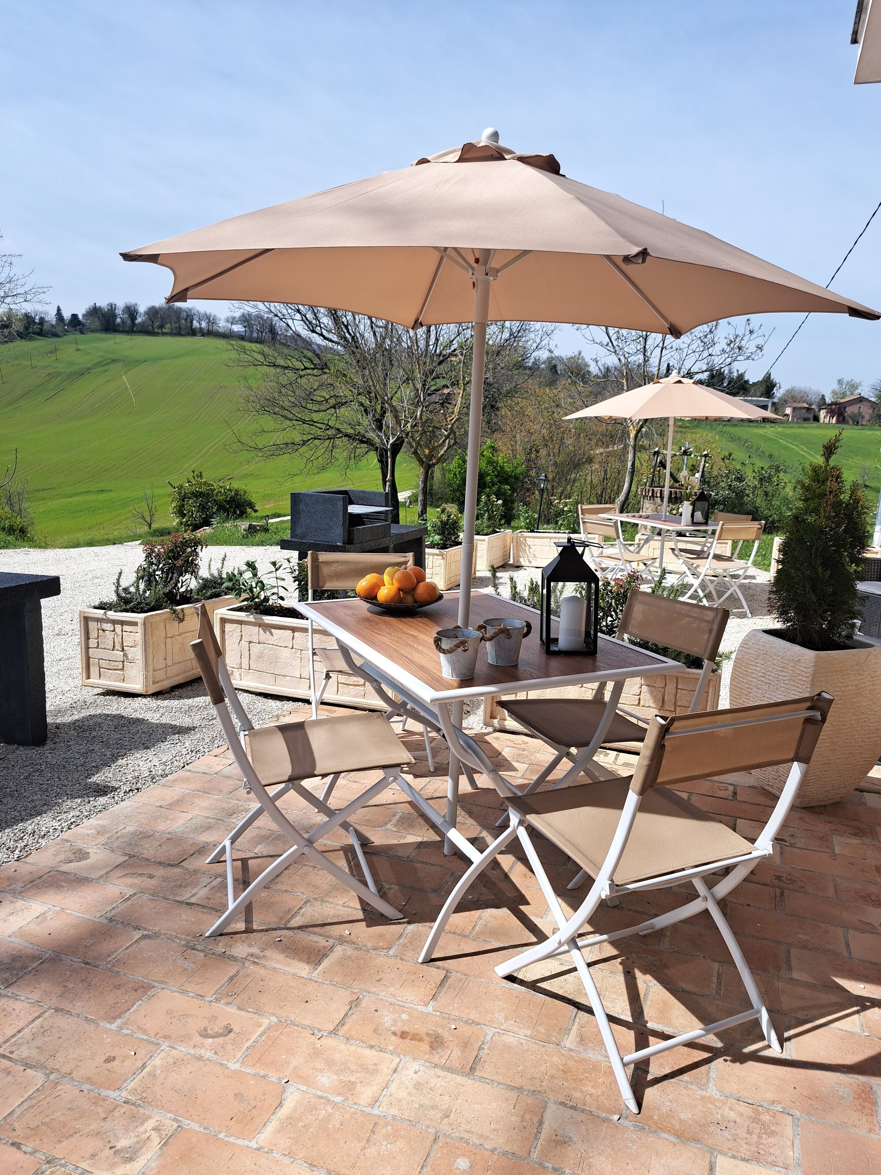 An outdoor patio with a table and chairs under a large umbrella, surrounded by planters and a view of a green field.