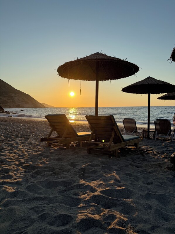 A beach with wooden lounge chairs and thatched umbrellas under a sunset.
