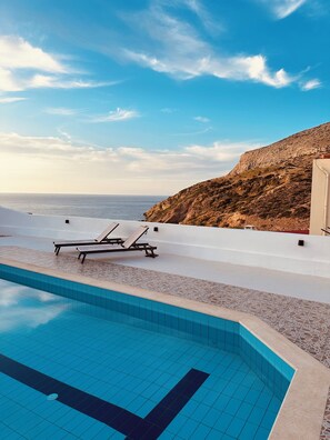 A pool area with lounge chairs and a view of the sea and a rocky hillside.