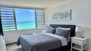 A bedroom with a large window, a bed with grey bedding, a nightstand with a lamp, and a wall art piece.