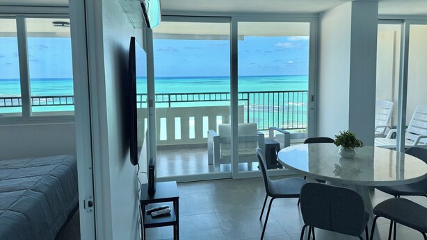 A room with a balcony overlooking a beach, a round dining table with chairs, a TV on a stand, and a bed with a blue cover.