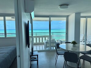 Una habitación con balcón con vista a la playa, una mesa de comedor redonda con sillas, un televisor sobre un mueble bajo y una cama con cubrecama azul.