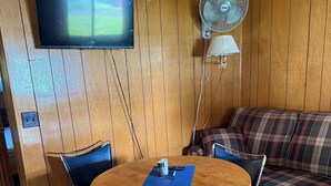 A room with a wooden paneled wall, a television mounted above a wooden table with chairs, a floor lamp, and a plaid sofa.