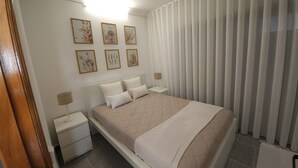 A bedroom with a bed, two bedside tables, a nightstand, a wall-mounted lamp, and a window with vertical blinds.
