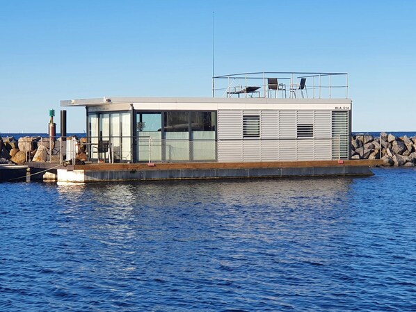 A modern floating house with a glass facade, a deck with chairs, and a view of the water. - Hausboot in Laboe an der Kieler Foerde (Laboe)