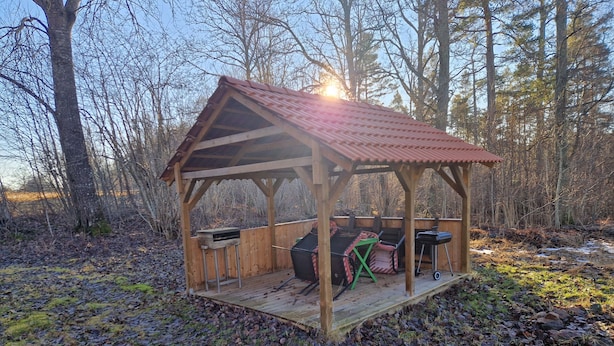 A wooden gazebo with a metal roof, a barbecue grill, and a table with chairs. - Vacation apartment Fagerhult (Kalmar län)
