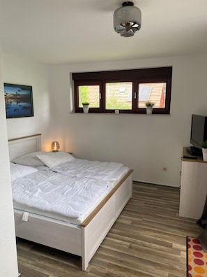 A bedroom with a bed, a bedside lamp, a TV, a window with plants, and a painting on the wall.