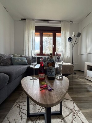 A living room with a sofa, a coffee table with bottles and glasses, a lamp, and a view through a window.