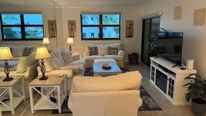 Un salon moderne avec des meubles blancs, un téléviseur à écran plat et de grandes fenêtres donnant sur des palmiers et des immeubles.