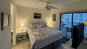Une chambre avec un lit, deux lampes de chevet, un ventilateur de plafond, une télévision et un balcon offrant une vue sur les bateaux et les bâtiments.