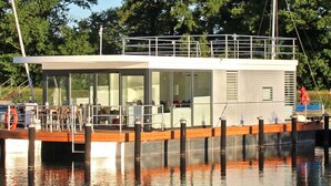 Ein modernes Hausboot an einem Steg, mit einer überdachten Deckterrasse und großen Fenstern.