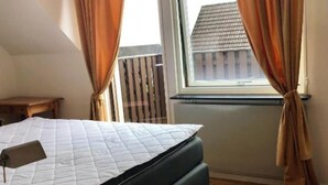 Une chambre à coucher avec un lit, une fenêtre avec des rideaux, un plancher en bois et une vue sur un bâtiment à l’extérieur.
