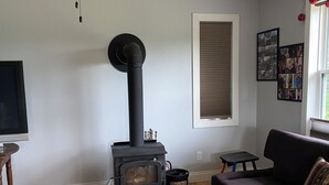 Un salon avec un poêle à bois noir, un ventilateur de plafond, un canapé noir et un plancher en bois.