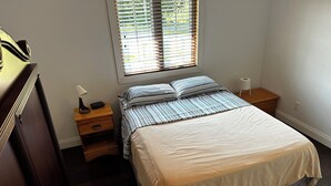 Une chambre à coucher comprenant un lit, une table de chevet, une commode, une fenêtre avec des stores et une lampe.